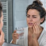 Woman in a white robe winces in pain while holding a glass of ice water and touching her cheek in a bathroom mirror.