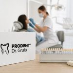 Dental shade guide and Prodent Dr. Gruia logo on a wooden table with a blurred dentist treating a patient in a bright, modern clinic.